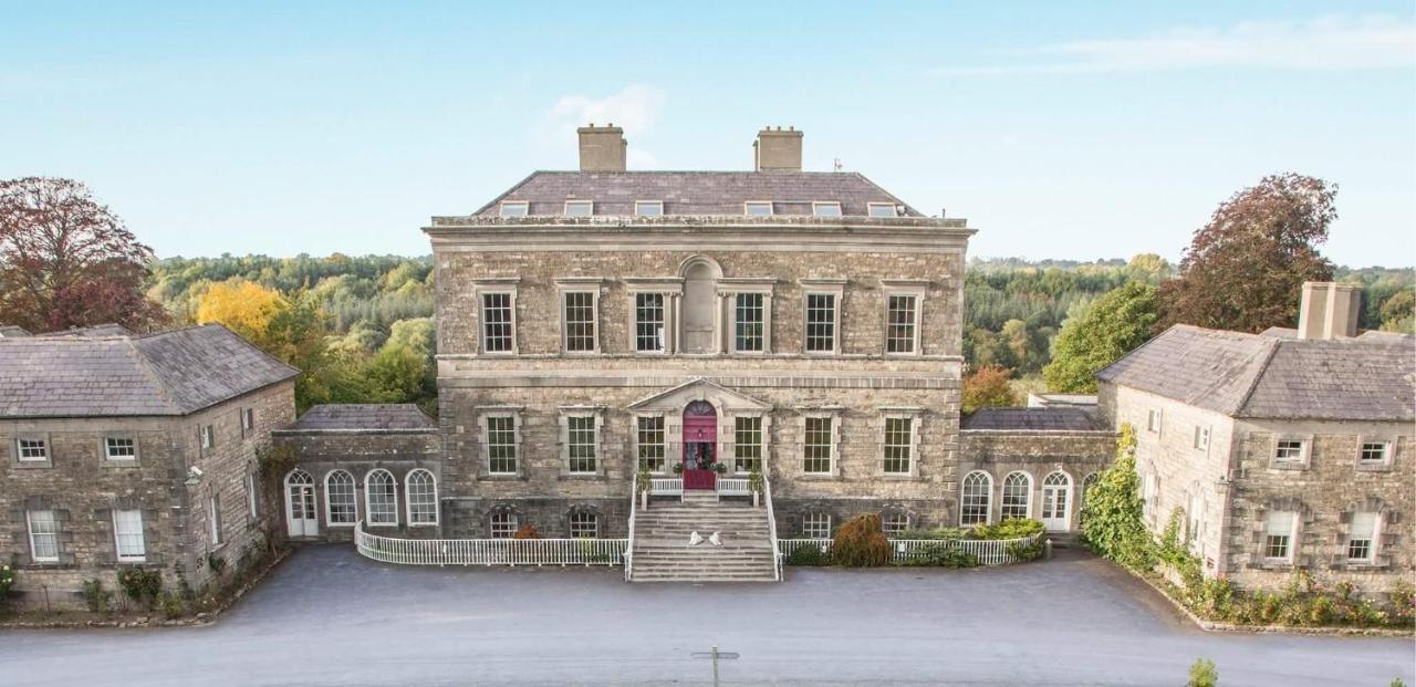 Historic stone mansion with symmetrical architecture, central red door, flanked by trees and greenery.