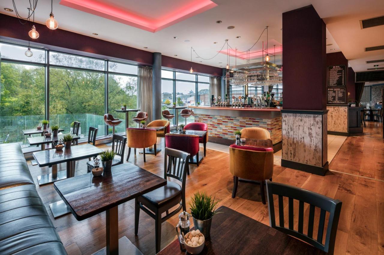 Modern restaurant interior with bar area, eclectic furniture, and large windows overlooking greenery.