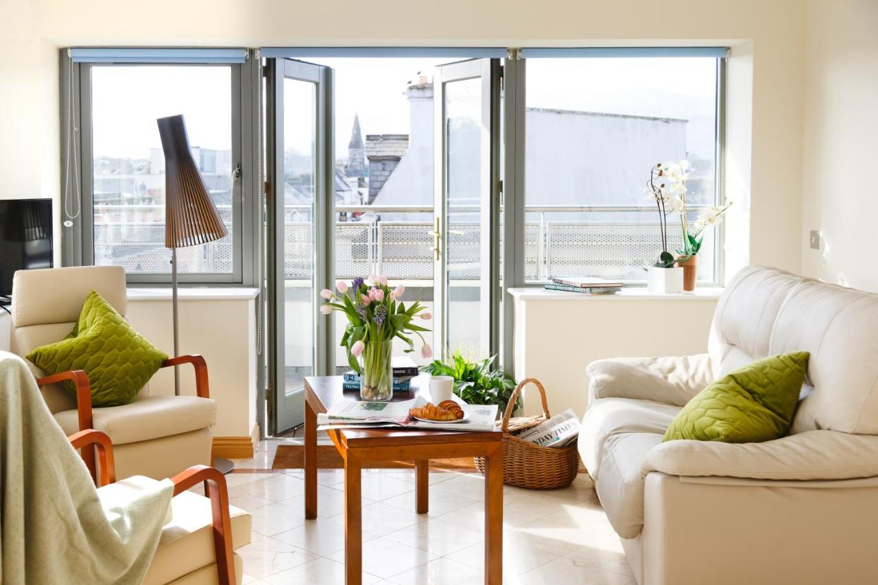 Bright living room with open balcony door, cozy seating, and a wooden coffee table adorned with flowers and pastries.