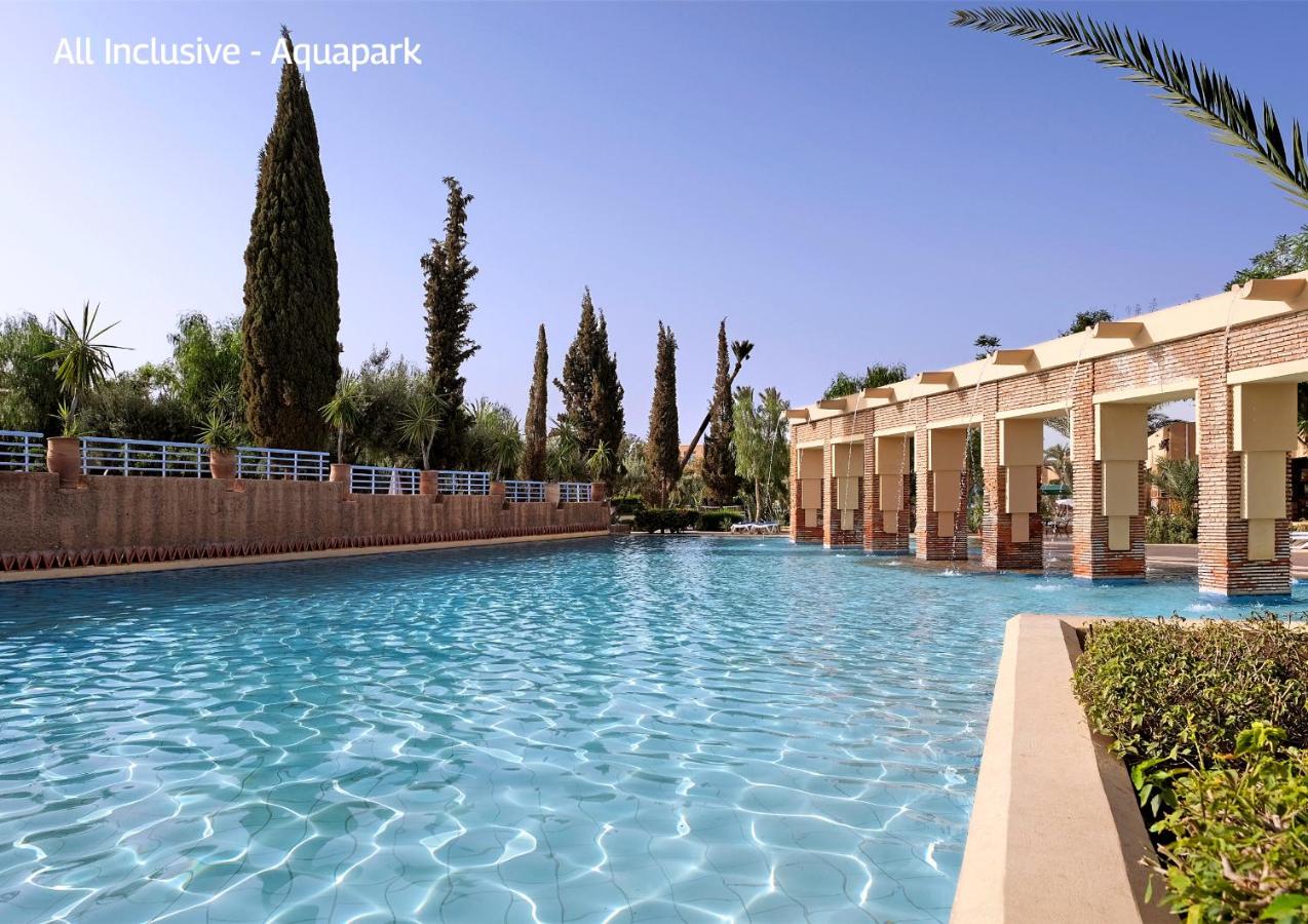 A large, tranquil swimming pool surrounded by tall trees and sunlit columns at an aquapark resort.